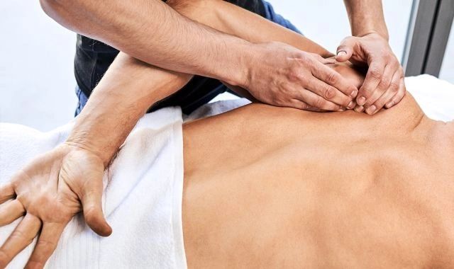 A therapist's hands and forearms working across a client's upper back and shoulder, a white towel folded at the lower back.